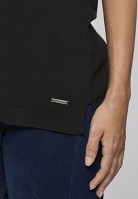 Svart stickad tröja med sidofickor, med en silverfärgad logotagg. Kombinerad med mörka denimbyxor, som visar en närbild av materialet och passformen.