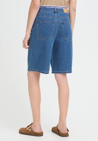 Shorts en jean bleu moyen avec une coupe décontractée, taille mi-haute, deux poches arrière et un ourlet brut. Chaussures plates en daim marron.