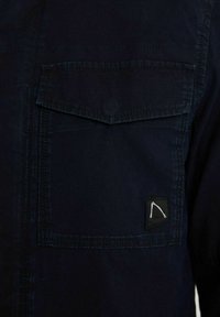 Navy blue shirt with a textured surface, featuring a chest pocket and a small logo patch in the corner. Stitching is visible along the seams.
