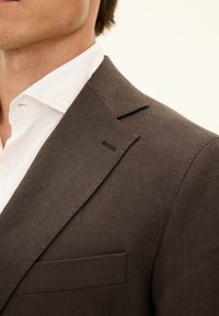 Blazer in lino marrone con colletto a scollo, tessuto testurizzato e una tasca frontale; indossato sopra una camicia bianca con colletto.