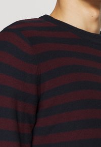 Personne portant un pull en tricot à rayures horizontales bordeaux et noires, montrant la zone des épaules et du cou.