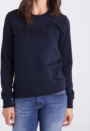 Femme portant un sweat-shirt bleu marine avec "TOMMY" embossé sur la poitrine et un jean bleu, debout devant un fond blanc uni.