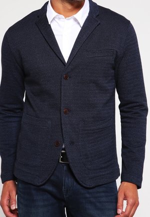 Hombre con blazer azul oscuro texturizado sobre una camisa blanca y jeans oscuros, con botones marrones en el blazer y un cinturón negro.