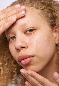 Primo piano di un volto con pelle marrone chiaro, occhi verdi e capelli ricci. La pelle ha una texture naturale con alcune imperfezioni; le mani sono appoggiate sulla fronte.