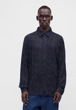 Jeune homme portant une chemise bleu foncé boutonnée avec un motif chevron et un jean bleu sur un fond clair uni.