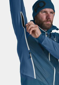 Men's blue athletic jacket with a zippered arm pocket, sleek design, textured patterns, and adjustable hood. Knit hat with logo detail.