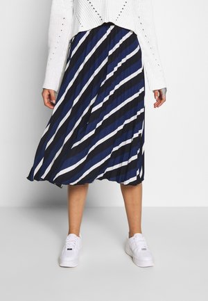 Femme portant une jupe plissée à rayures diagonales bleues, noires et blanches, un pull en maille blanc et des baskets blanches, debout devant un fond uni.