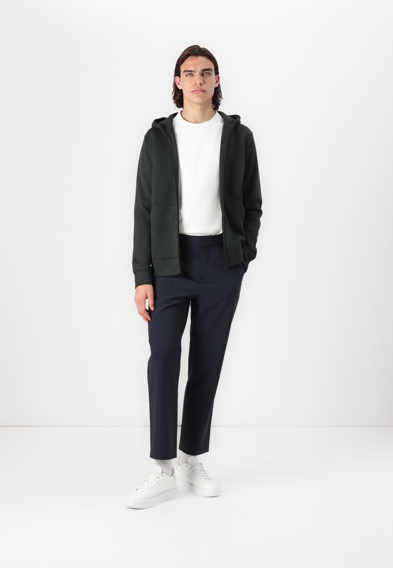 Zielona bluza z kapturem, biała koszulka z okrągłym dekoltem, granatowe spodnie i białe sneakersy. Strój ma luźny krój z gładkimi teksturami i minimalistycznym designem.