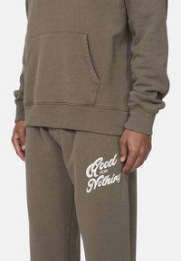 Brązowy sweatshirt z kieszenią z przodu, w po�łączeniu z dopasowanymi spodniami dresowymi z białym napisem "Good for Nothing" na udzie. Miękka faktura.