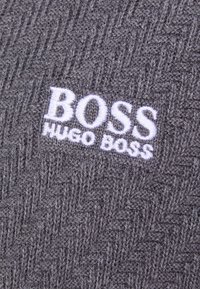 Šedá pletená látka s herringbonovým vzorem. Obsahuje bílou vyšívanou nášivku "BOSS HUGO BOSS" ve spodní části.