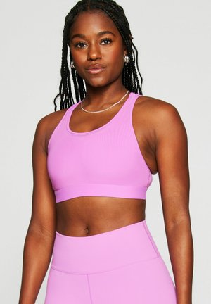 Athletic woman with braided hair wearing matching light purple sports bra and high-waisted leggings, posing against a plain background.