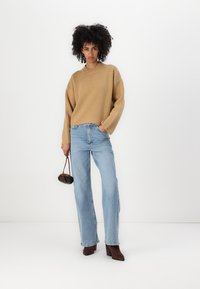 Maglione a maglia marrone chiaro con scollatura a coste, abbinato a jeans dritti a vita alta di un blu sbiadito. La modella tiene una borsa piccola.