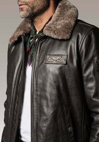 Veste bomber en cuir marron avec un col en fausse fourrure, des poches latérales et un patch avec l'inscription "American Aircraft Academy". Fermeture éclair visible.