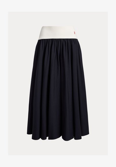 Φούστα μέχρι το γόνατο από σκούρο μπλε ύφασμα με λευκή ελαστική ζώνη στη μέση. Διαθέτει πιετέ σχέδιο και ένα μικρό κόκκινο λογότυπο ως λεπτομέρεια.