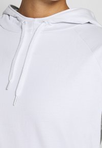 Gros plan d'une personne portant un sweat à capuche blanc avec des cordons visibles et l'encolure sur un fond uni.
