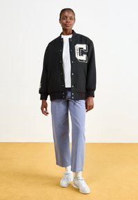 Giacca bomber nera con bottoni a pressione frontali, logo "C" ricamato, abbinata a una maglietta bianca, pantaloni azzurri chiari e sneakers bianche.