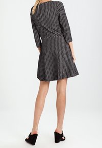 Robe noire à manches trois-quarts, avec un motif à pois, une jupe évasée et un corsage ajusté. Chaussures à talons hauts noires.