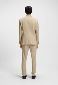 Man standing facing away, wearing a beige suit with black shoes, against a plain light gray background.