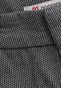 Nahaufnahme eines schwarz-weißen Hahnentrittmusters mit einer umgeschlagenen Kante und einem teilweise sichtbaren weißen Etikett mit rotem und schwarzem Text.