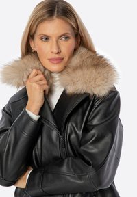 Giubbotto di pelle nera con un voluminoso colletto in pelliccia sintetica beige. Presenta una chiusura con zip, maniche lunghe e dettagli di cucitura visibili.