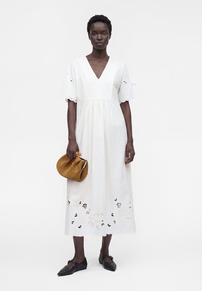 Femme portant une robe midi blanche avec des manches à festons et un ourlet festonné, tenant une pochette tissée marron et des ballerines noires pointues.