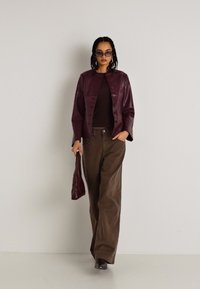 Burgundská kožená bunda cez hnedý top, v kombinácii s širokými hnedými nohavicami a zodpovedajúcou taškou. Modelka so tmavými slnečnými okuliarmi.