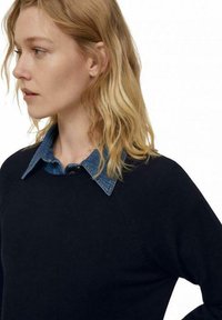 Marineblauer Pullover mit strukturierter Ripp-Textur, über einem verblassten Denim-Hemd mit spitzem Kragen getragen. Das Design umfasst Raglanärmel.