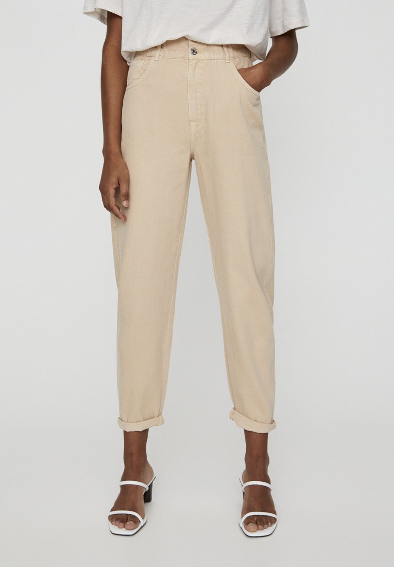 Osoba ubrana w beżowe spodnie z wysokim stanem z podwiniętymi nogawkami, białe sandały na obcasie z paskami oraz w wpuszczoną w spodnie jasnobeżową koszulkę z krótkim rękawem.