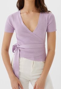 Kvinna i en ribbad lavendelfärgad wrap-top med korta ärmar som är knuten i midjan, kombinerat med högmidjade off-white byxor.