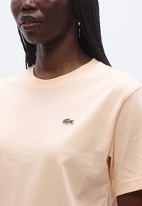 Lyse ferskenfarvet bomuld T-shirt med rund hals og korte ærmer. Har en lille grøn krokodille-logo broderet på brystet. Glat tekstur.