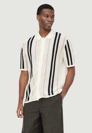 Muž nosiaci krátky rukáv, zapínanú pletenú košeľu s vertikálnymi čierno-bielymi pruhmi, v tmavých nohaviciach, stojaci s jednou rukou v vrecku.