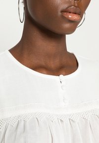 Blusa bianca con scollo rotondo, dettaglio in pizzo testurizzato e tre bottoni rivestiti in tessuto sul davanti. Materiale leggero e liscio.
