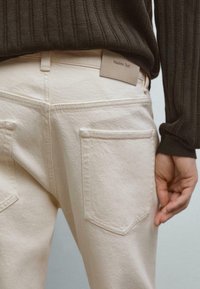 Beige Denim-Jeans mit Gesäßtaschen und einem Massimo Dutti-Label, getragen mit einem dunklen, gerippten Pullover, wobei eine Hand entspannt an der Seite hängt.