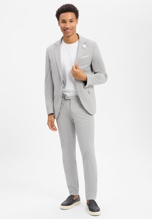 Junger Mann lächelnd, trägt hellgrauen Anzug mit weißem Hemd und schwarzen Slippern, steht vor schlichtem weißem Hintergrund.