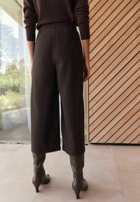 Braune weit geschnittene Hosen aus strukturiertem Stoff mit zwei Gesäßtaschen und hohem Bund, kombiniert mit olivgrünen High-Heels.