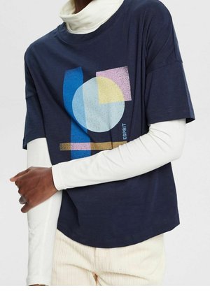 Henkilö, jolla yllään laivastonsininen graafinen t-paita, jossa abstrakteja geometrisia muotoja sinisen, keltaisen, vaaleanpunaisen ja valkoisen sävyissä, sekä alla pidettävä pitkähihainen paita.