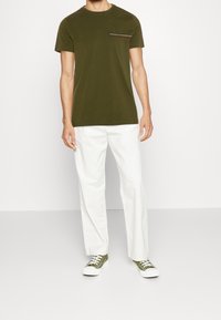 Olivenfarvet kortærmet t-shirt med rund hals og brystlomme. Parret med hvide løstsiddende bukser og grønne sneakers.