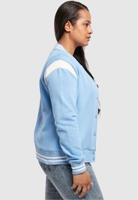 Giacca varsity blu chiaro con chiusura a bottoni, spalle bianche e polsini a righe, realizzata in tessuto liscio. Vista laterale mostrata.