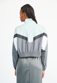 Geacă de vânt Nike în albastru deschis și gri, cu un design scurtat, talie elastică și accente negre contrastante. Textură de material neted.