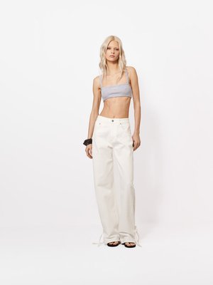 Kõhn blond naine, kellel on hall crop-top, valged laia sääreosaga püksid paeltega ääres, must käevõru ja mustad sandaalid, seisab valge tausta ees.