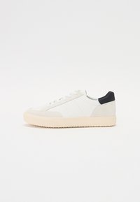 White leather sneaker with a textured suede toe, beige rubber sole, and a black heel accent. Features lace-up design and side panel stitching.