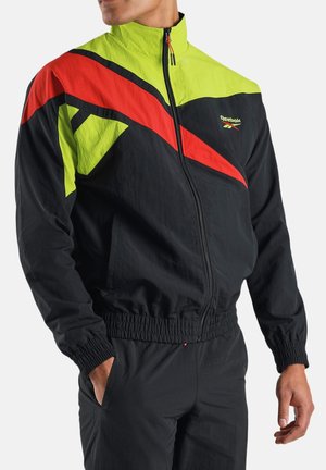 Uomo con tuta Reebok nera con strisce diagonali giallo neon e rosse, in piedi con una mano in tasca contro uno sfondo semplice.