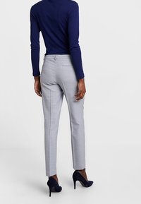 Světle šedé kalhoty na míru s kostkovaným vzorem, v kombinaci s tmavě modrým topem s dlouhým rukávem a tmavými podpatky, z pohledu zezadu.