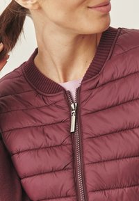 Giacca imbottita bordeaux con design trapuntato, colletto a costine e chiusura centrale con zip. I dettagli delle cuciture aggiungono texture; il tessuto appare liscio.