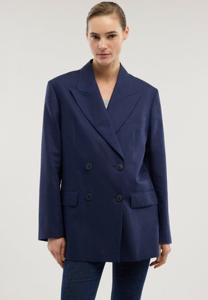 Donna che indossa un blazer doppiopetto blu navy con ampi revers sopra una maglietta grigia e jeans scuri, in piedi davanti a uno sfondo chiaro e uniforme.