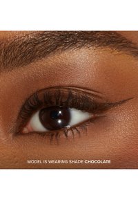 Close-up van een oog met een bruine iris, dikke wimpers en gedefinieerde eyeliner met vleugels. Huidskleur is diep met een gladde structuur. Opvallende makeupkleur: Chocolade.