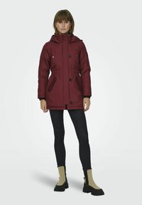 Parka isolante bordeaux avec capuche, dotée d'une taille à cordon de serrage, de deux poches avant et de finitions noires. Le modèle porte un jean noir et des bottines.