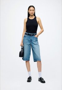 Vrouw die een zwarte mouwloze top draagt, knielange blauwe denimshorts, witte sokken, zwarte loafers en een kleine zwarte handtas vasthoudt.