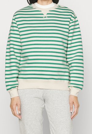 Personne portant un sweat-shirt à rayures horizontales vertes et blanches avec des poignets crème et un pantalon de survêtement gris, debout devant un fond uni.