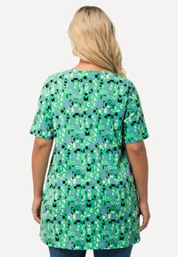 Vihreä tunika, jossa on geometrinen kuvio mustilla ja vaaleanpunaisilla yksityiskohdilla, pehmeästä kankaasta valmistettu, rento malli, lyhyet hihat ja pyöreä pääntie.